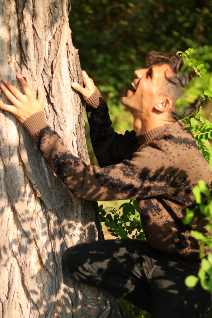 Eine Person kniet im Wald an einem großen Baumstamm, hält sich mit beiden Händen fest und wirkt konzentriert im Kontakt mit der Natur