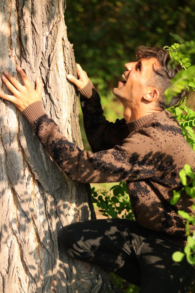 Eine Person kniet im Wald an einem großen Baumstamm, hält sich mit beiden Händen fest und wirkt konzentriert im Kontakt mit der Natur